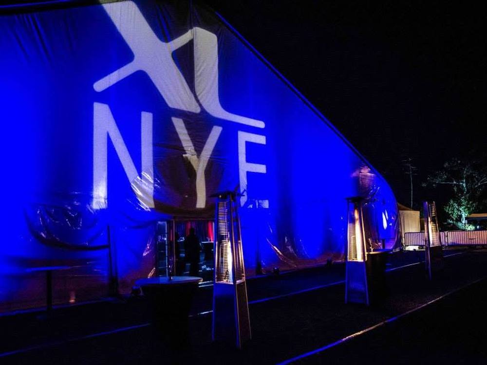 The XL NYE tent exterior at night, lit blue with the event logo projected across the fabric.