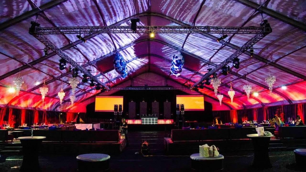 The XL NYE tent at Newport Dunes before doors: truss, chandeliers, stage and VIP seating in an empty room.